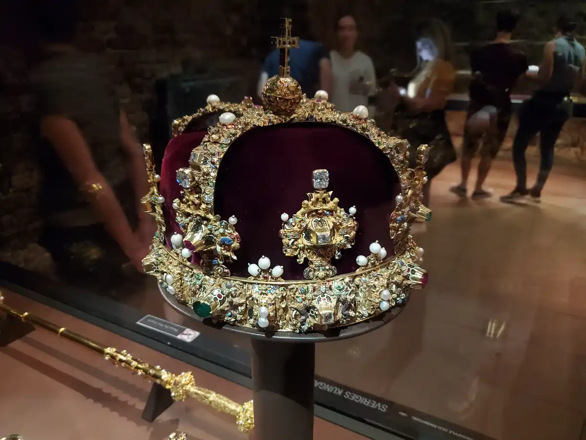 A heavily jeweled, ornate golden crown with pearls and gemstones sits on a display stand inside a museum, with blurred…