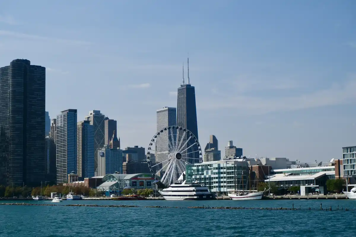 A scenic photo of the Chicago skyline, featuring the Willis Tower, a large Ferris wheel, and boats on the water. The…