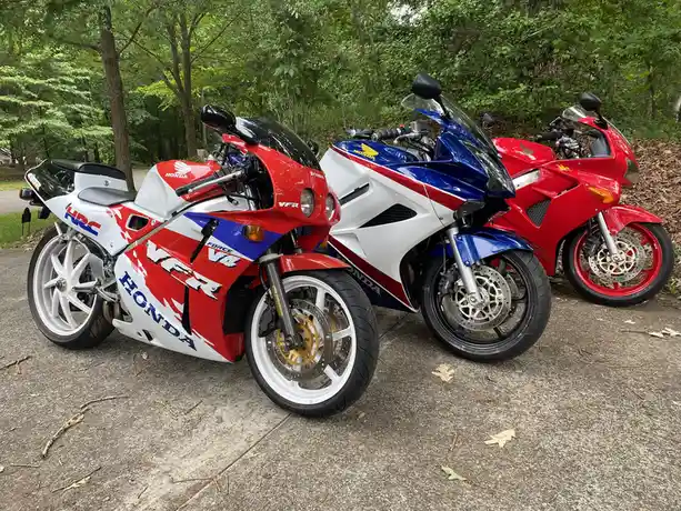 Three Honda sport motorcycles are parked side-by-side on a paved surface in front of a wooded area. The bike on the left is…