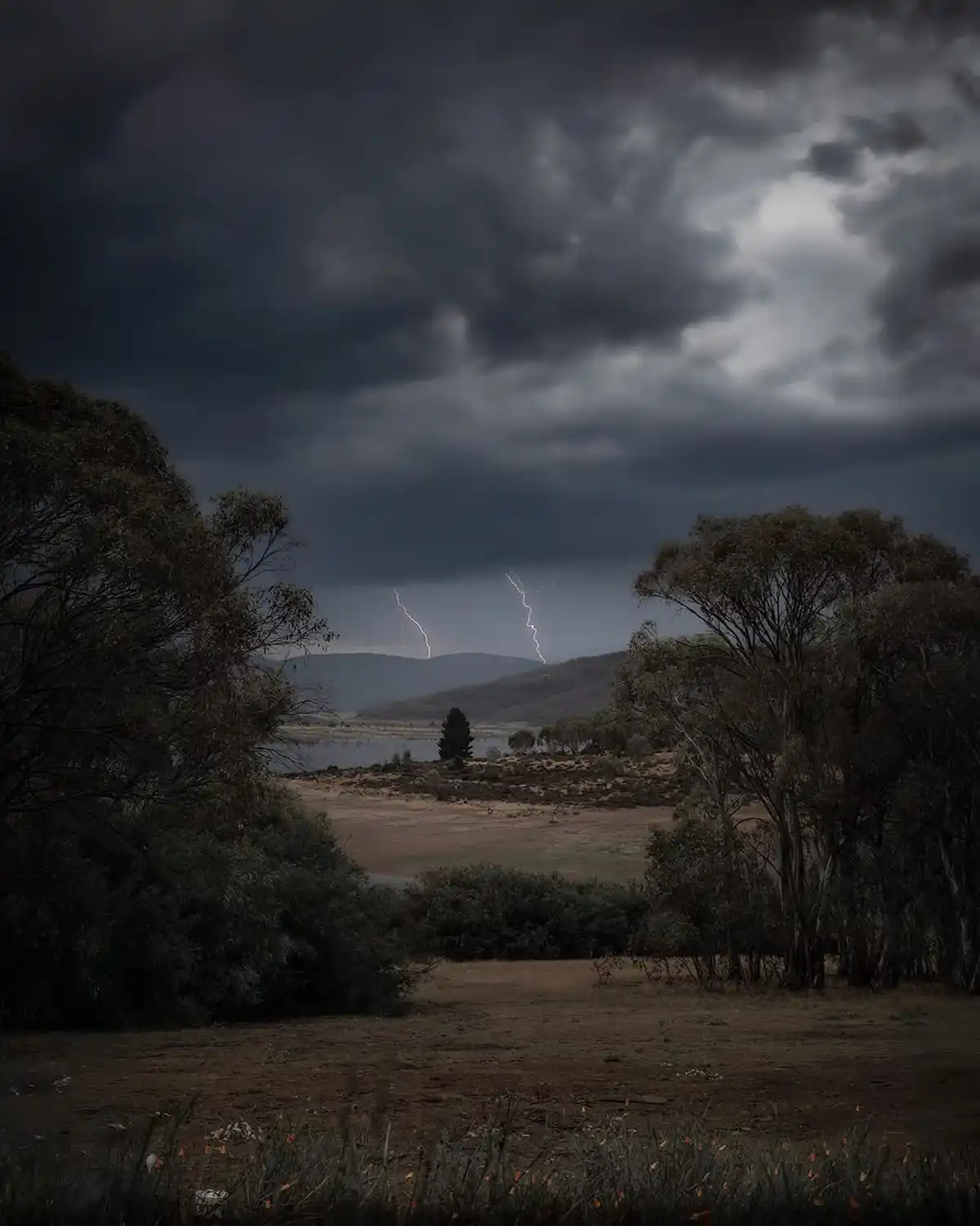 A dark, moody landscape photo shows a stormy sky with two bright lightning bolts striking over distant hills, framed by…