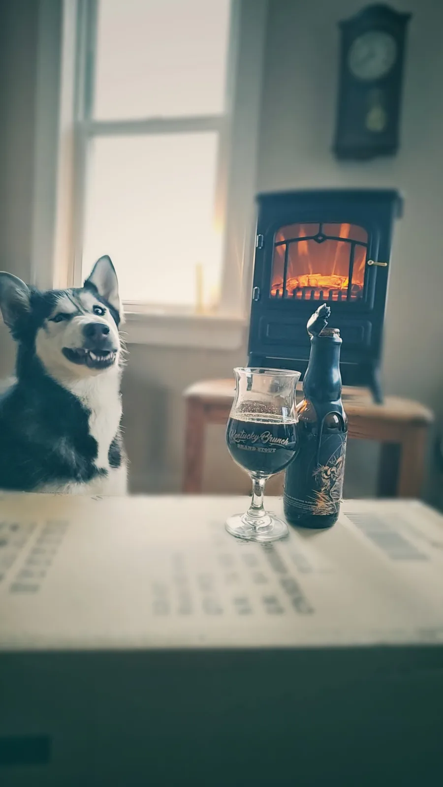 A black-and-white husky dog sits at a table next to a glass and bottle of 'Kentucky Brunch Brand Stout' beer, with a lit…