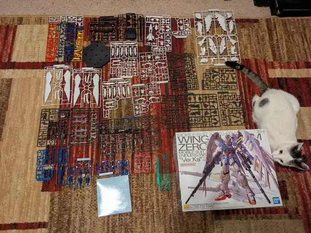 An overhead shot shows numerous plastic sprues from a Gundam model kit spread across a patterned rug, with a white and gray…