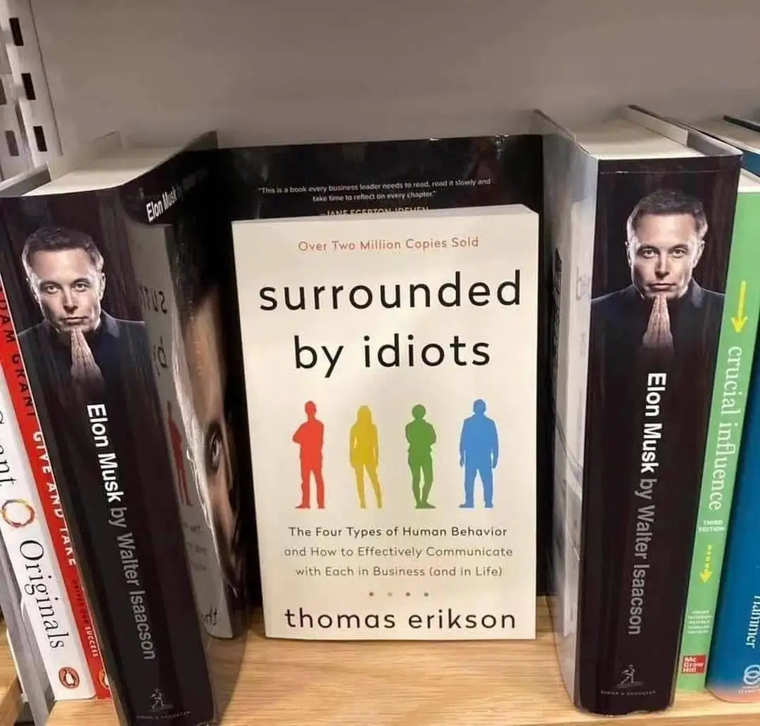 A bookshelf displays a book titled 'surrounded by idiots' by Thomas Erikson, flanked on both sides by books about Elon Musk…