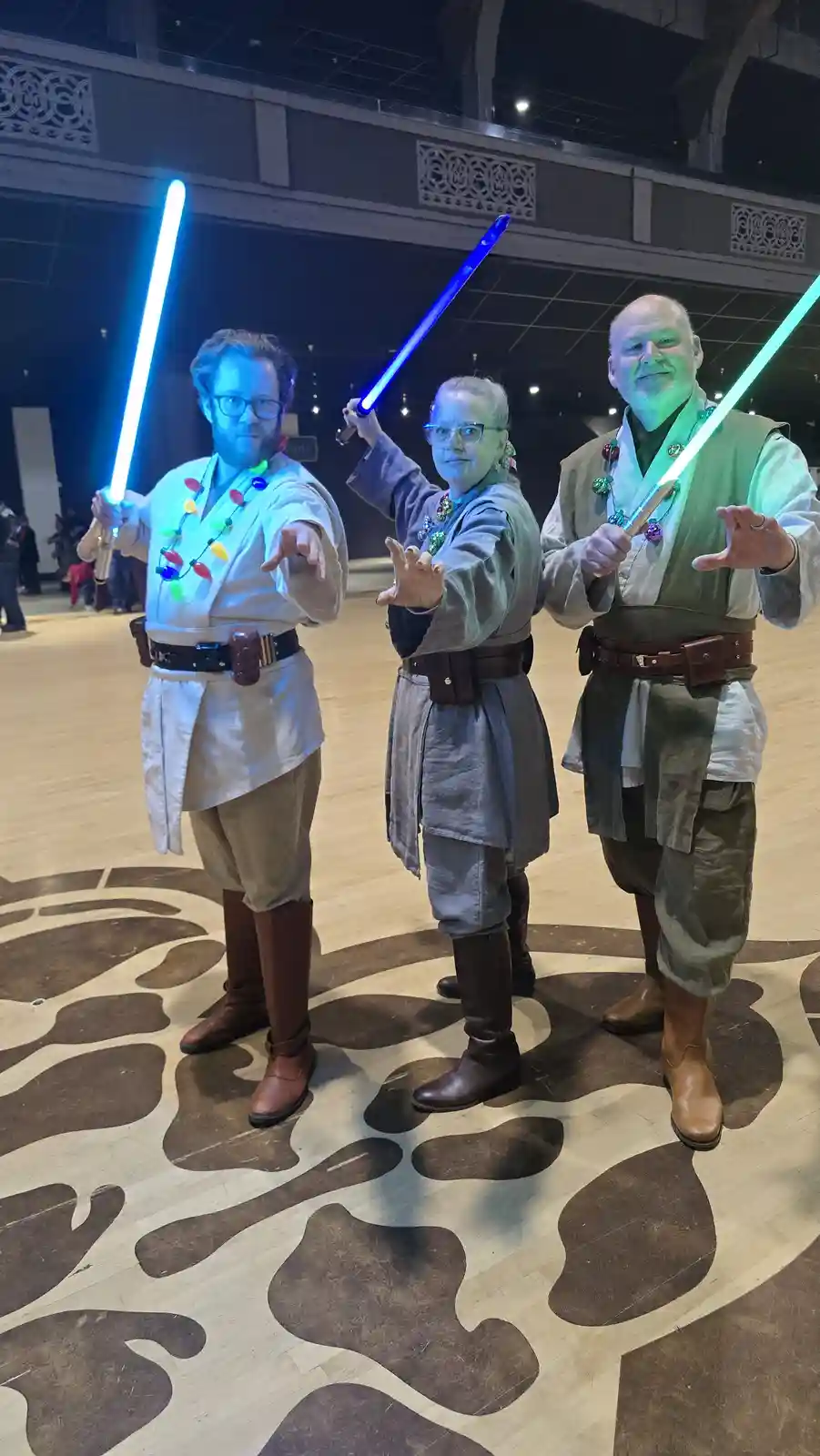 Three people dressed as Star Wars Jedi, holding glowing lightsabers, are posing with festive Christmas lights strung around…