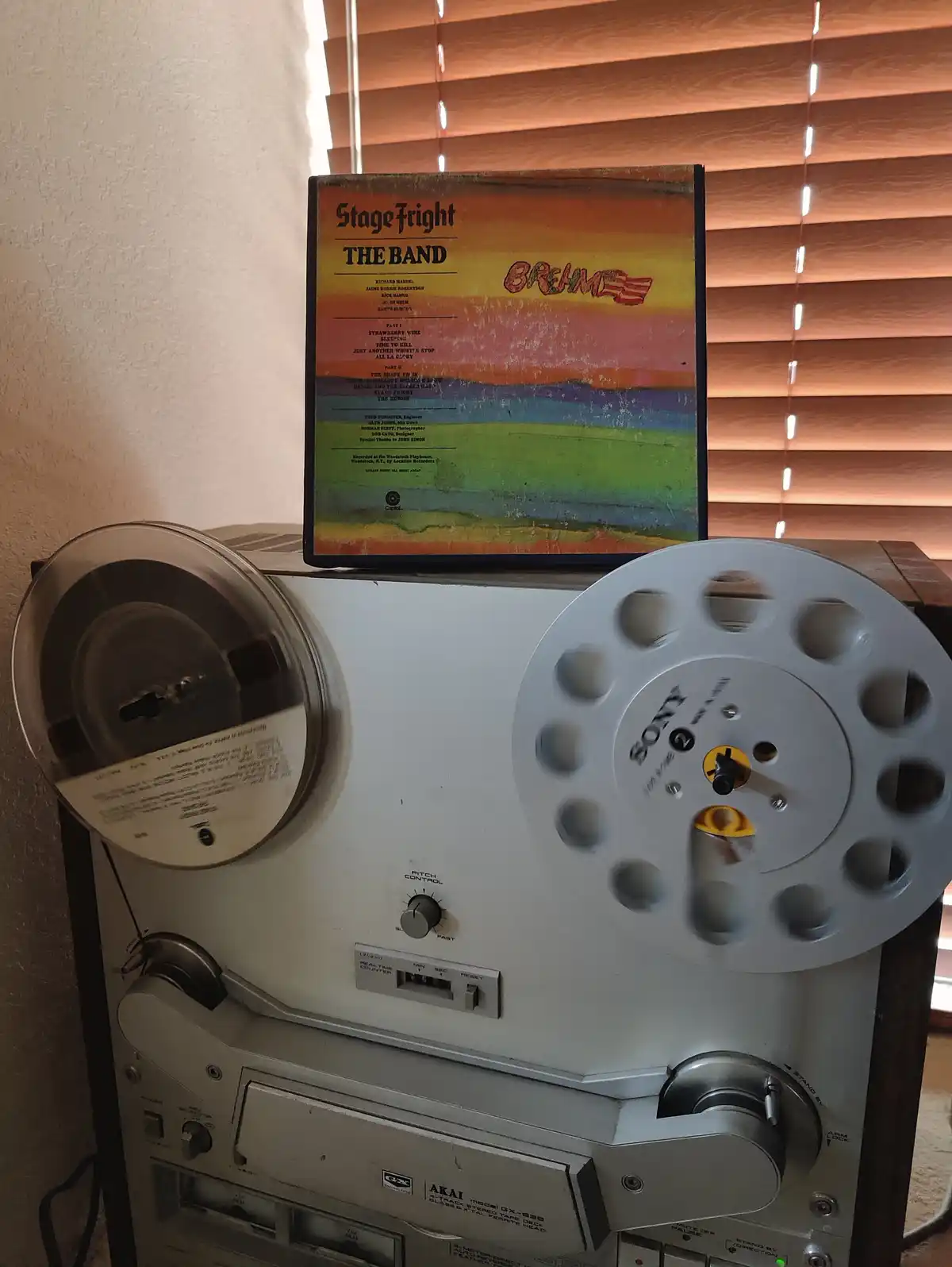 A vintage Akai reel-to-reel tape recorder with a Sony reel sits on a wooden surface, with a worn album cover for 'Stage…