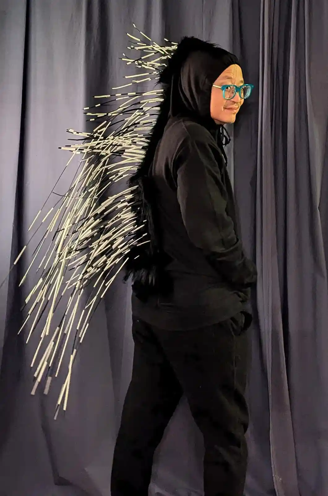 A man in a black hoodie and bright blue glasses stands sideways against a gray curtain, looking back over his shoulder with…