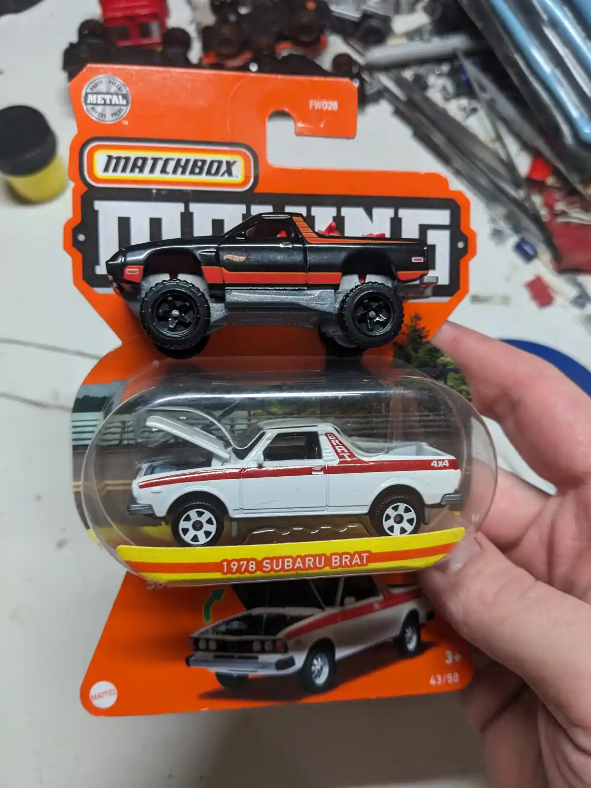 A hand holds a Matchbox toy car package featuring a white 1978 Subaru BRAT with red stripes and open hood, labeled '1978…