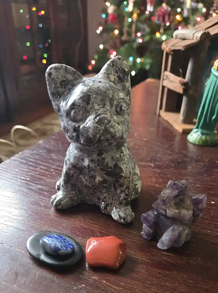 A stone carving of a French bulldog sits on a wooden table surrounded by various crystals including amethyst, lapis lazuli…