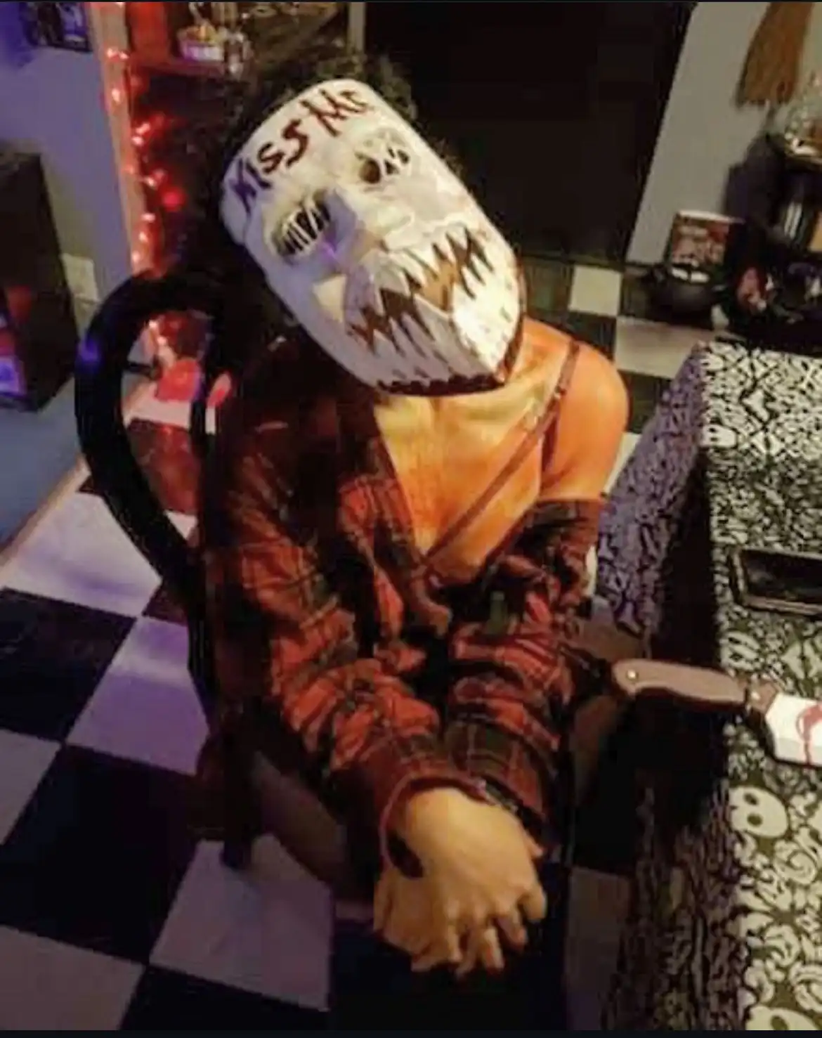 A person wearing a white, skull-faced mask with 'kiss me' written on the forehead sits slumped in a chair. They are dressed…
