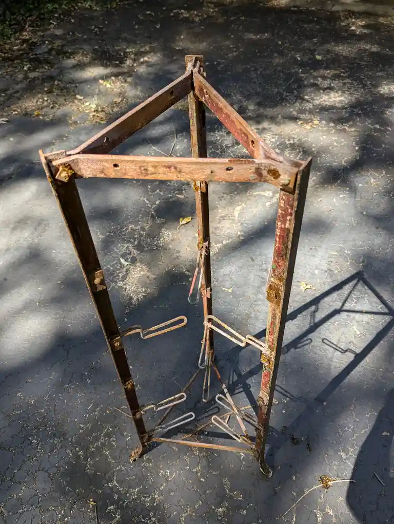 A rusty, weathered metal frame, likely from an old bicycle rack or similar structure, stands on cracked pavement outdoors…