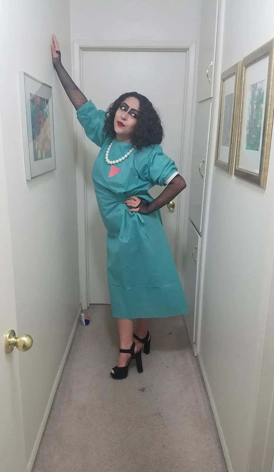 A person in a teal surgical gown with a pink triangle on the chest, black fishnet gloves, and a pearl necklace stands in a…