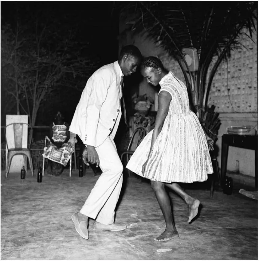 A black-and-white photo shows a man and a woman dancing together outdoors at night, both looking down with awkward…