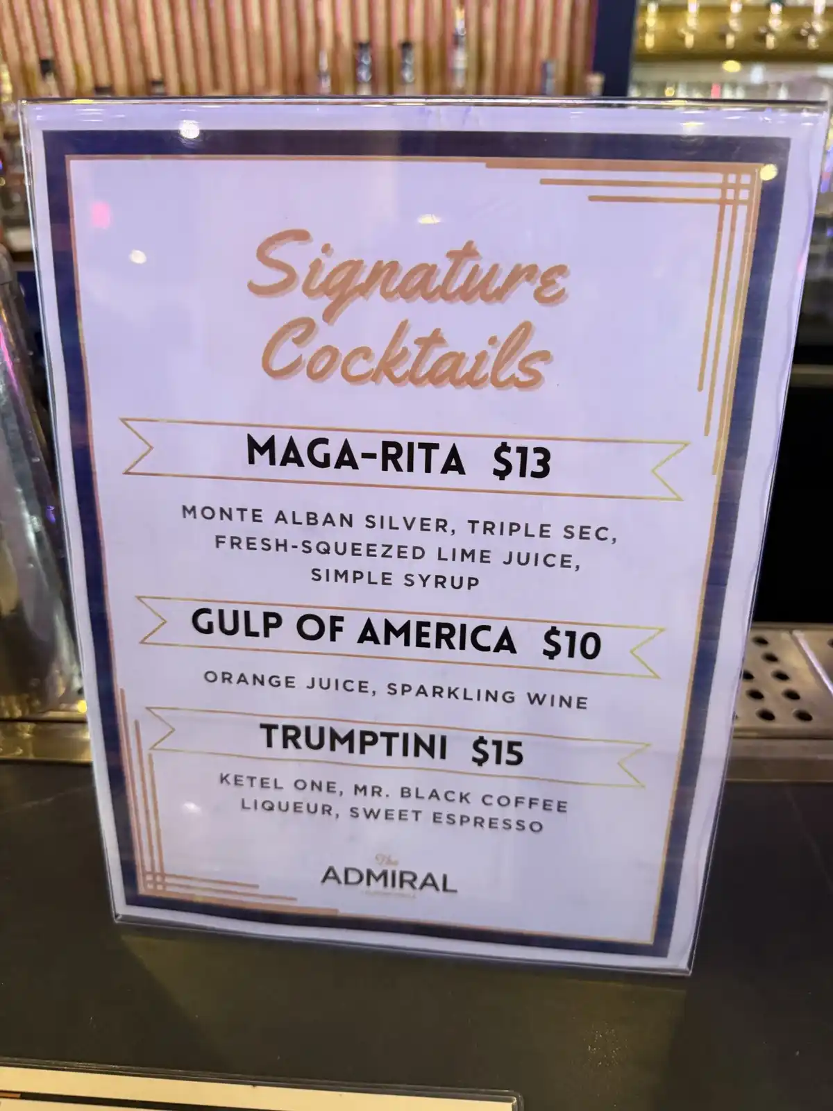 A bar menu titled 'Signature Cocktails' lists three drinks with politically themed names: 'MAGA-RITA' ($13), 'GULP OF…