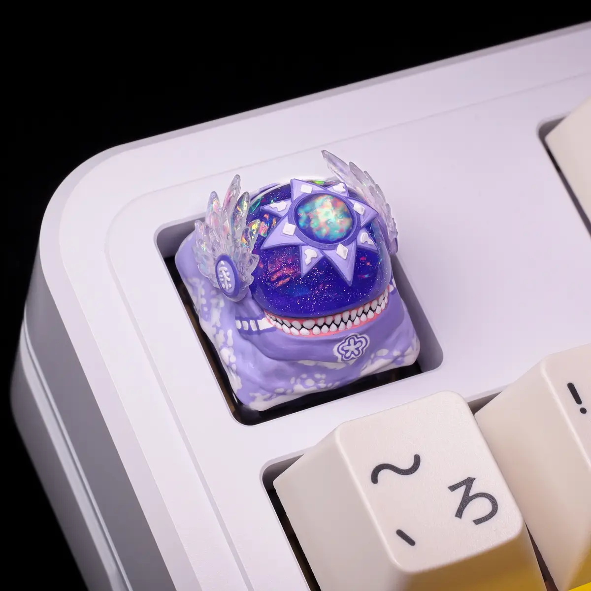 A close-up of a highly customized, ornate purple keyboard keycap shaped like a smiling skull with glittery galaxy patterns…