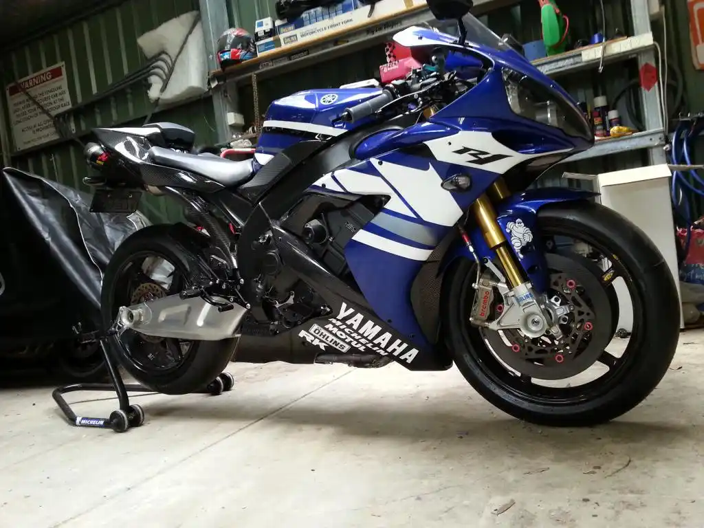 A blue and white Yamaha R1 sport motorcycle is parked on a stand in a cluttered garage, with shelves of tools and parts…