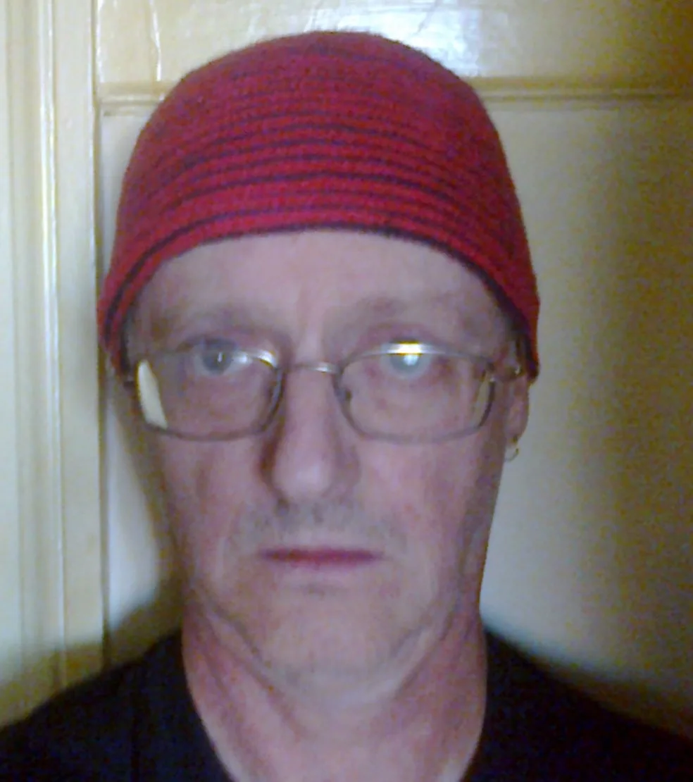 A close-up photo of a middle-aged man with a stern, unimpressed expression, wearing a red knit beanie and glasses, standing…
