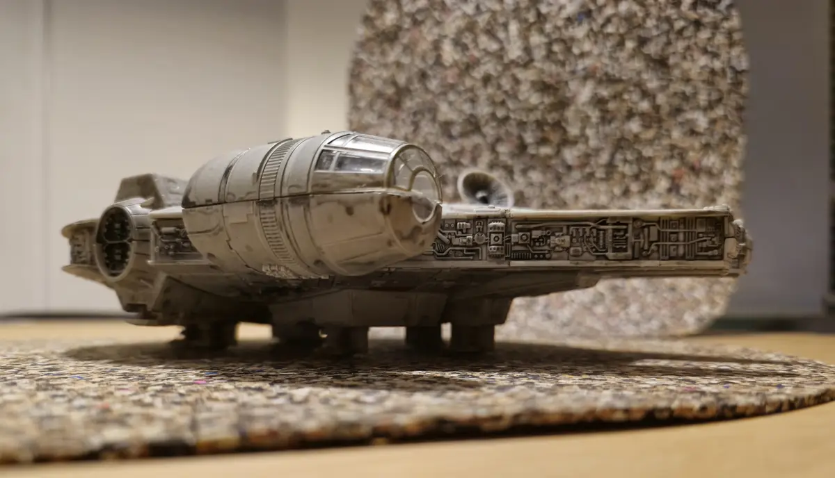 A detailed model of the Millennium Falcon spaceship sits on a textured rug, with a blurry background. The overlaid text…