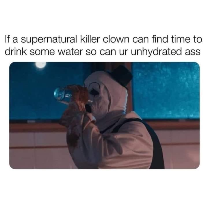 A person wearing a white clown costume with a black top hat and a painted-on smiley face mask is drinking from a clear…