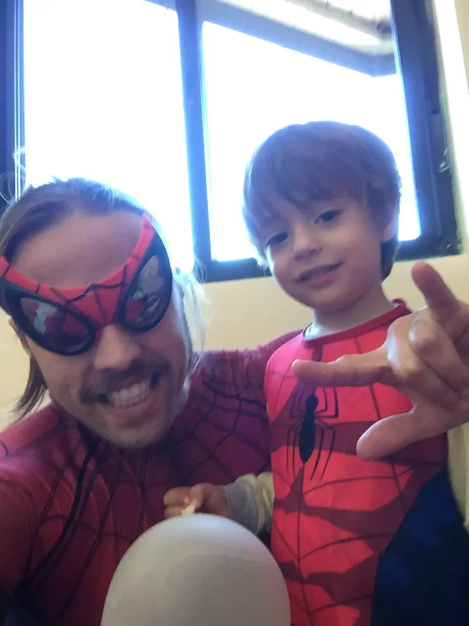 A man and a young boy, both dressed in red and blue Spider-Man costumes, pose together indoors near a window. The man wears…
