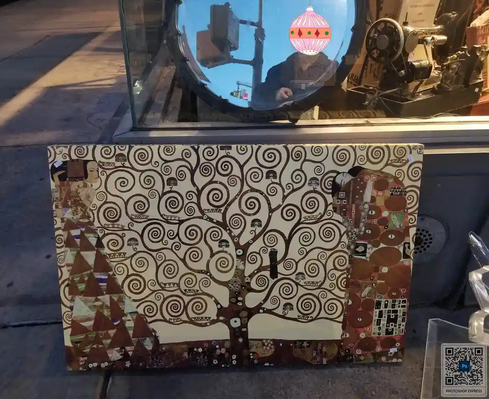 A canvas print of Gustav Klimt's 'The Tree of Life' is propped up outside a shop window, with a pink Christmas ornament…