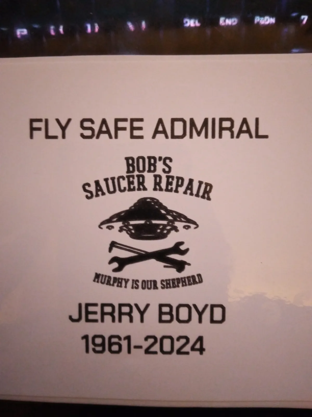 A printed memorial card for Jerry Boyd (1961-2024) features a UFO logo, crossed wrenches, and the text 'FLY SAFE ADMIRAL'…