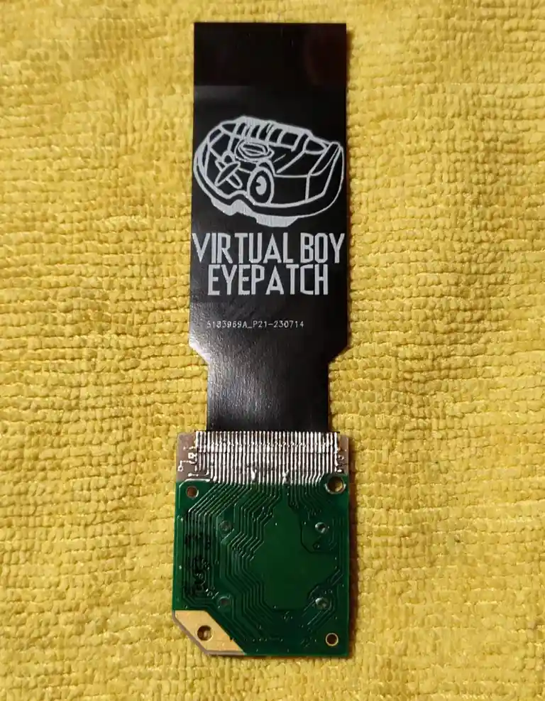 A close-up photo shows a black flexible circuit board with a white line drawing of a Virtual Boy headset and the text…