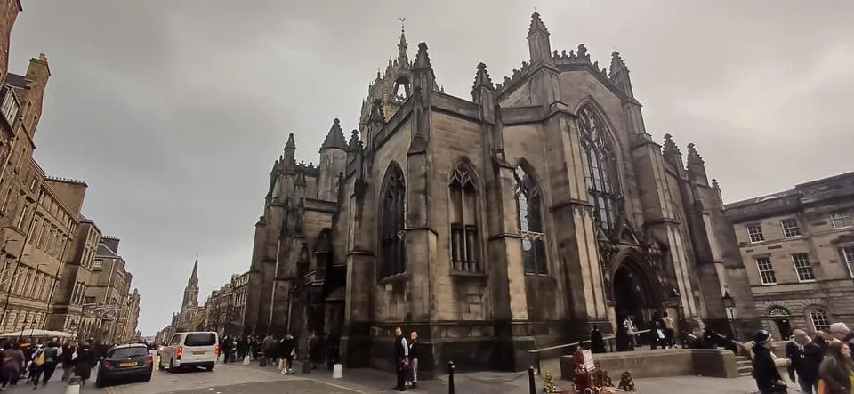 A wide-angle photo of a large, ornate gothic-style stone church with pointed spires and arched windows, situated on a busy…