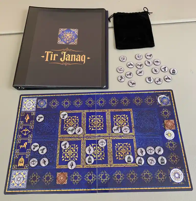 A tabletop game called 'Tir Janag' is displayed with its blue and gold patterned board, a black binder-style box, a velvet…