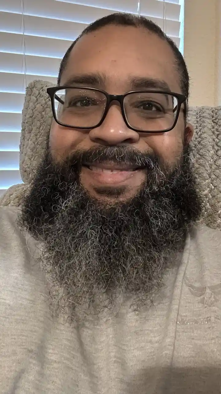 A close-up selfie of a smiling man with a thick, curly beard showing significant gray hairs, wearing black-rimmed glasses…