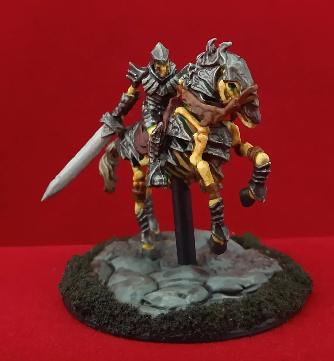 A painted miniature of a knight in full armor riding a skeletal horse, both adorned with metallic and bone-like details…
