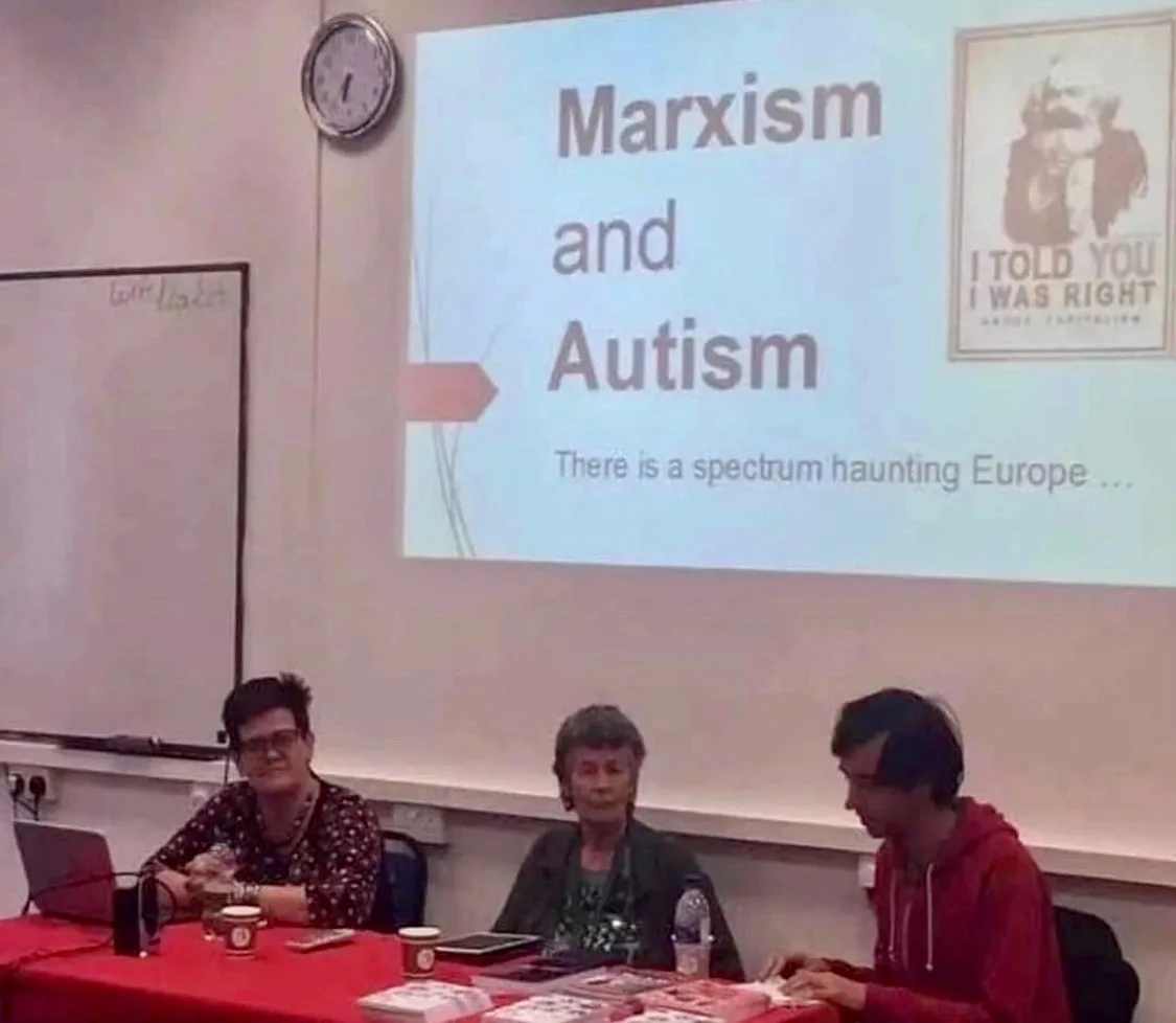 Three people sit at a table in front of a projector screen displaying a presentation titled 'Marxism and Autism' with the…