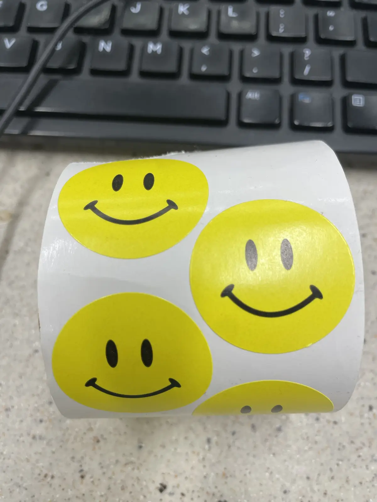 A roll of yellow smiley face stickers sits on a speckled countertop in front of a black keyboard. Three of the visible…