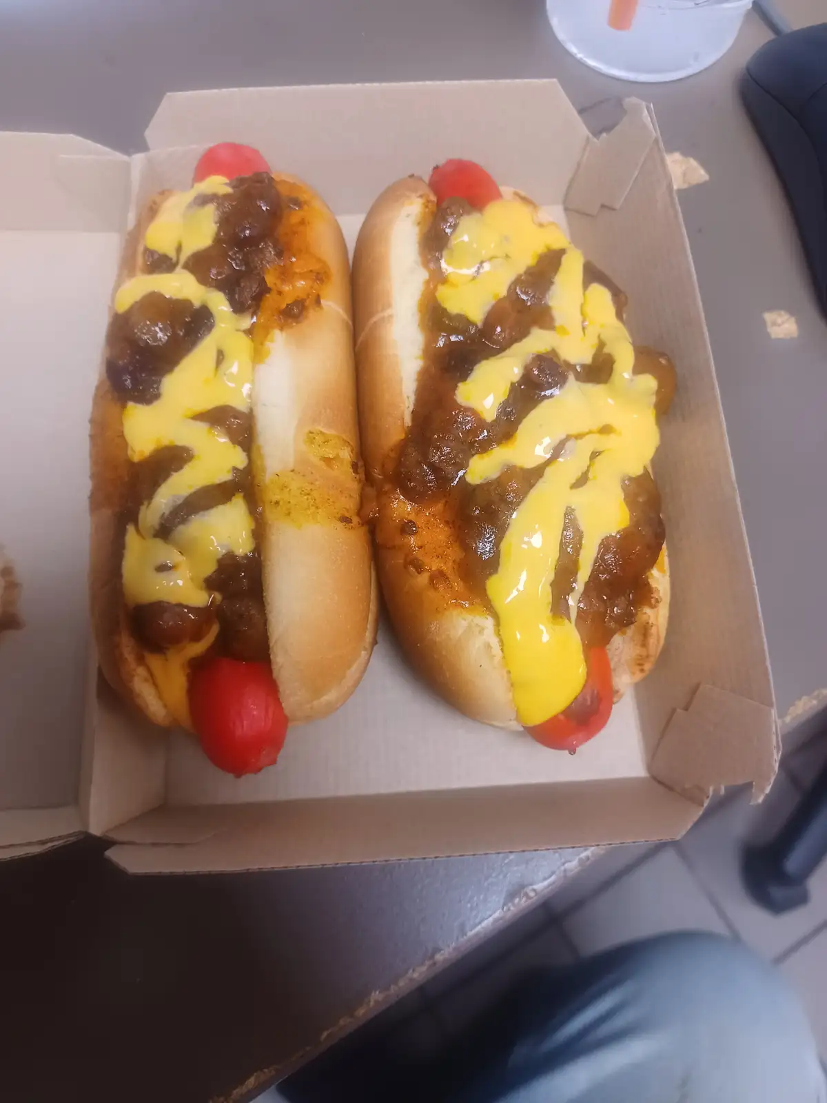 Two chili cheese hot dogs sit side-by-side in a cardboard tray, each topped with brown chili and bright yellow cheese sauce…