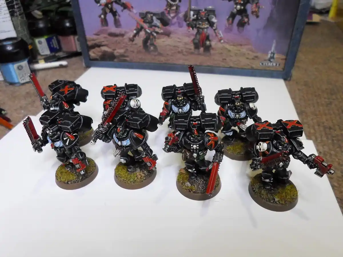 Five painted miniature warhammer 40k space marines in black armor with red accents and skulls, standing on grassy bases…