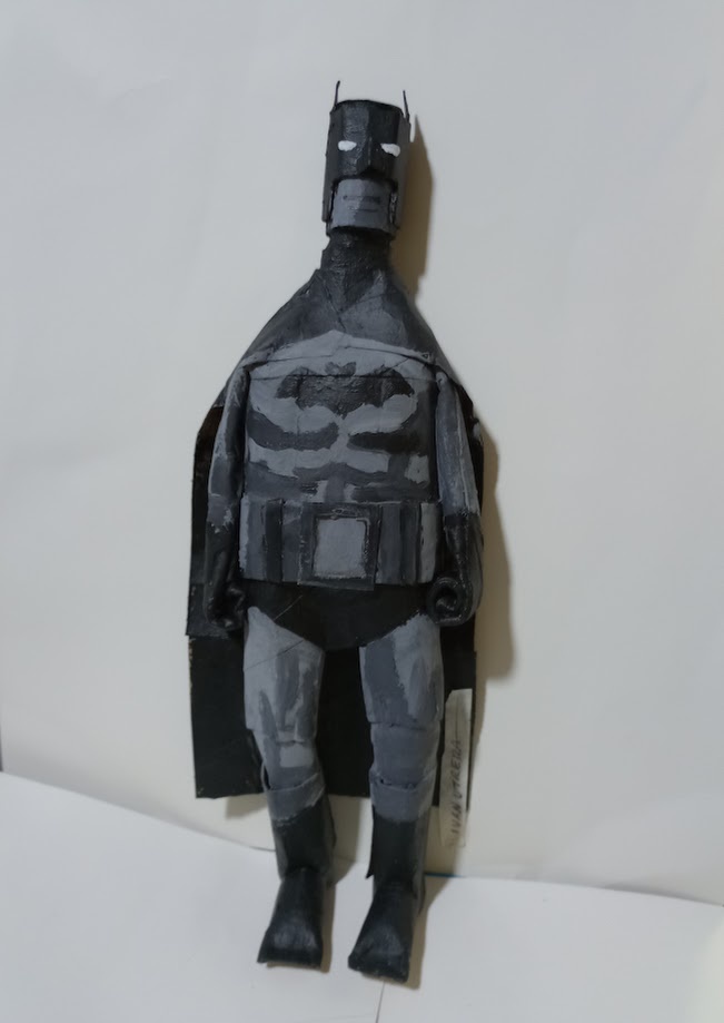 A handmade, crudely painted Batman figure made from cardboard or similar material stands against a plain white background…