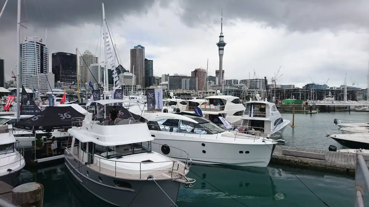 A harbor scene with numerous luxury yachts docked, set against a city skyline featuring the Sky Tower. The overlaid text…