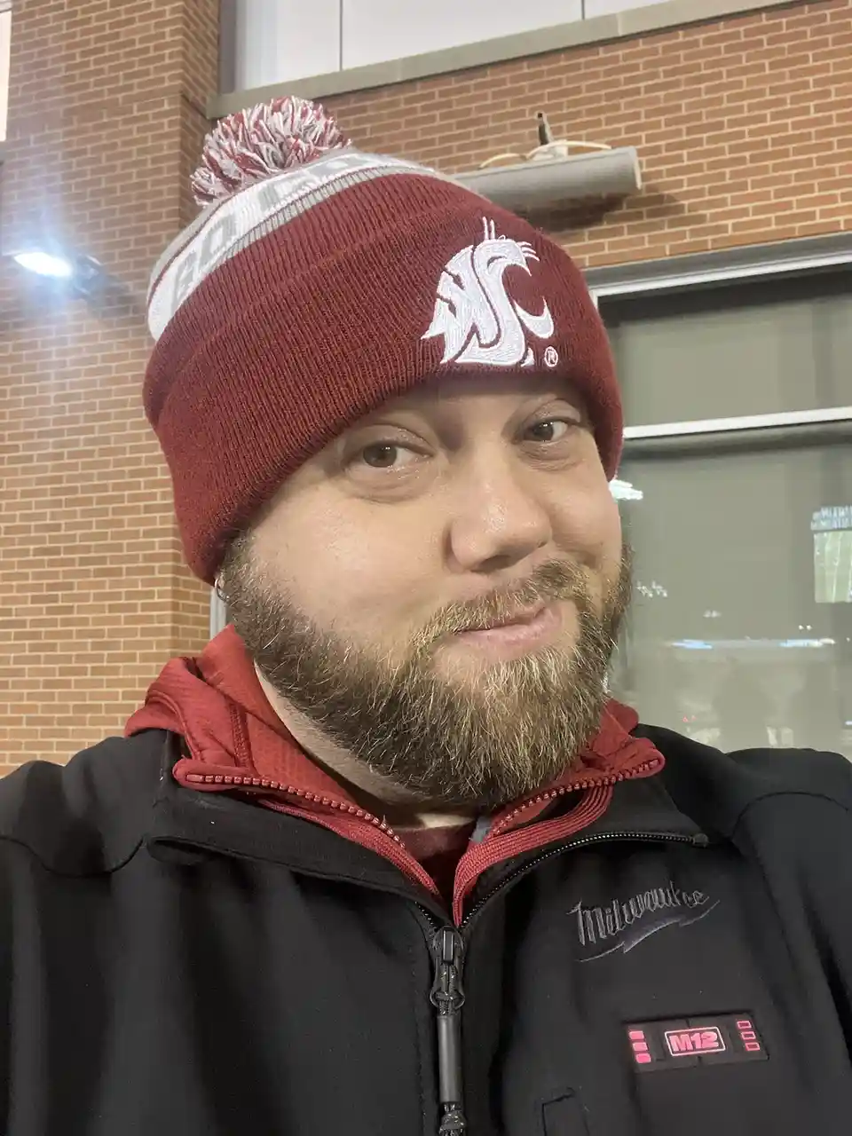 A man with a beard wearing a red and white Washington State Cougars beanie and a black Milwaukee M12 branded jacket poses…