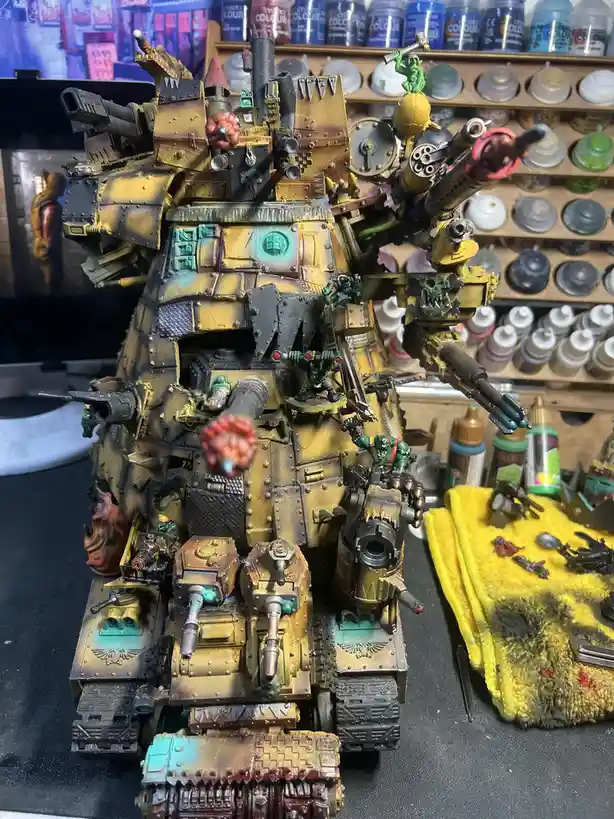 A heavily customized, multi-layered, and weapon-packed Warhammer 40k tank model sits on a workbench, surrounded by paint…