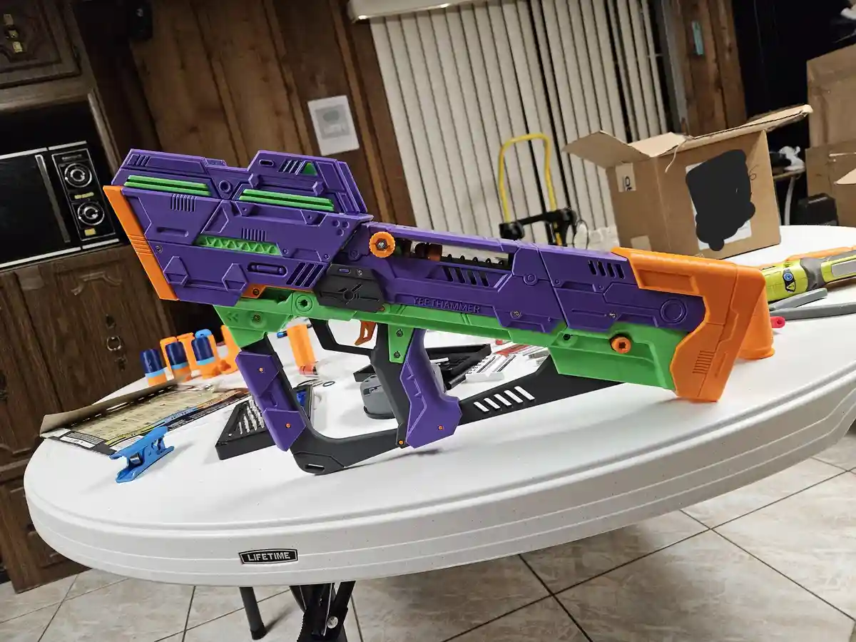 A large, custom-built toy blaster with purple, green, and orange plastic parts sits on a white Lifetime folding table. The…