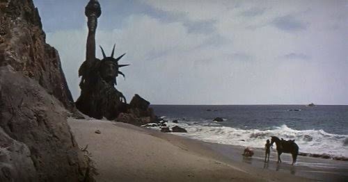 A desaturated, cinematic shot shows the Statue of Liberty partially submerged and broken on a rocky beach, with ocean waves…