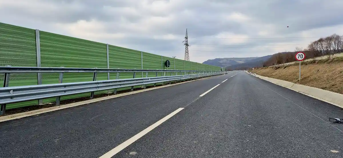 An empty asphalt road stretches forward under a cloudy sky, bordered by a long green noise barrier and a metal guardrail. A…