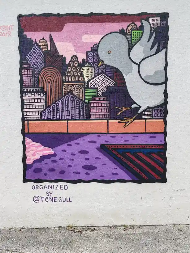 A colorful mural depicts a large white dove perched on a ledge overlooking a stylized cityscape with purple and pink tones…