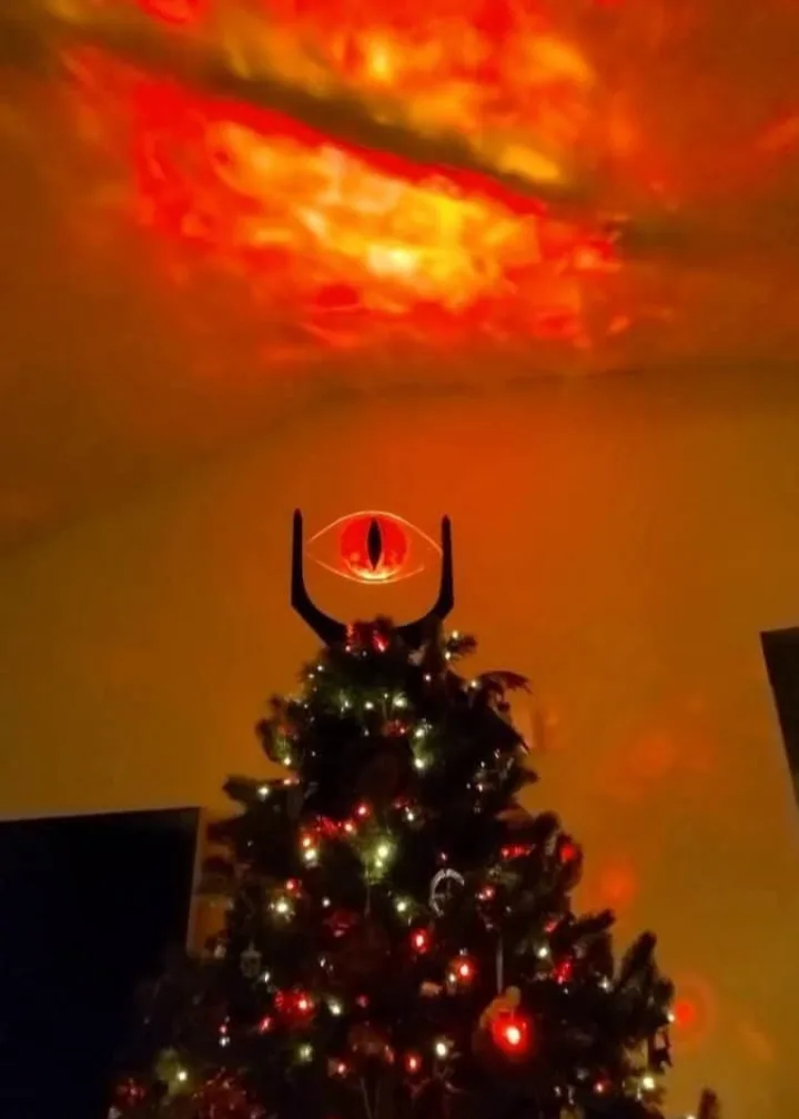 A decorated christmas tree with lights and ornaments has a glowing red eye of sauron projected onto its top, with a fiery…