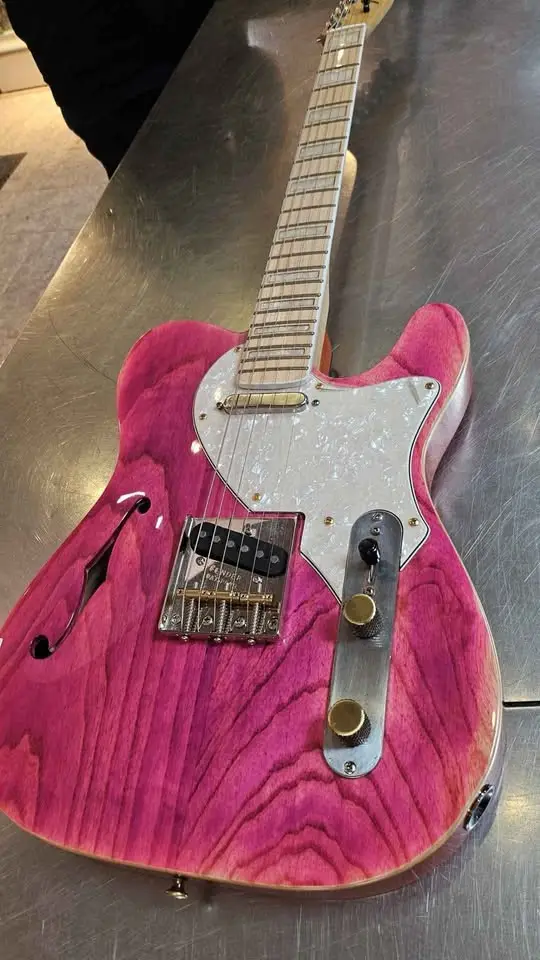 a vibrant pink electric guitar with a wood grain finish and white pickguard rests on a brushed metal surface. The overlaid…