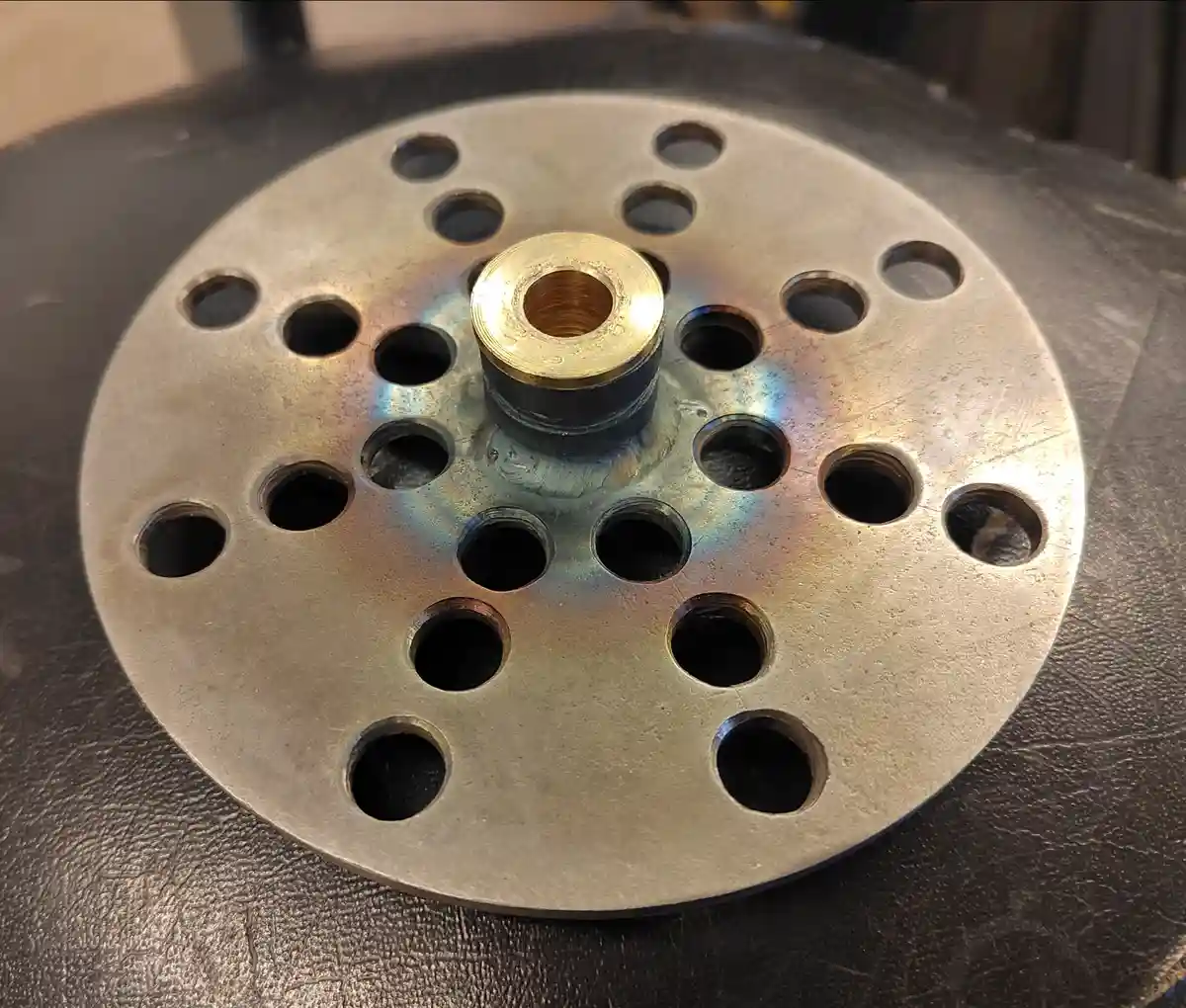 A close-up photo shows a circular metal plate with multiple evenly spaced holes and a central brass fitting, exhibiting a…