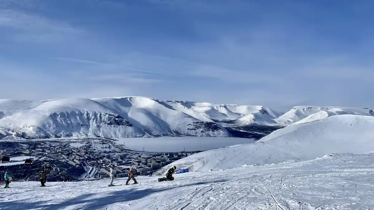 A wide snowy mountain landscape with skiers and snowboarders on a slope, overlooking a frozen lake and a town below under a…