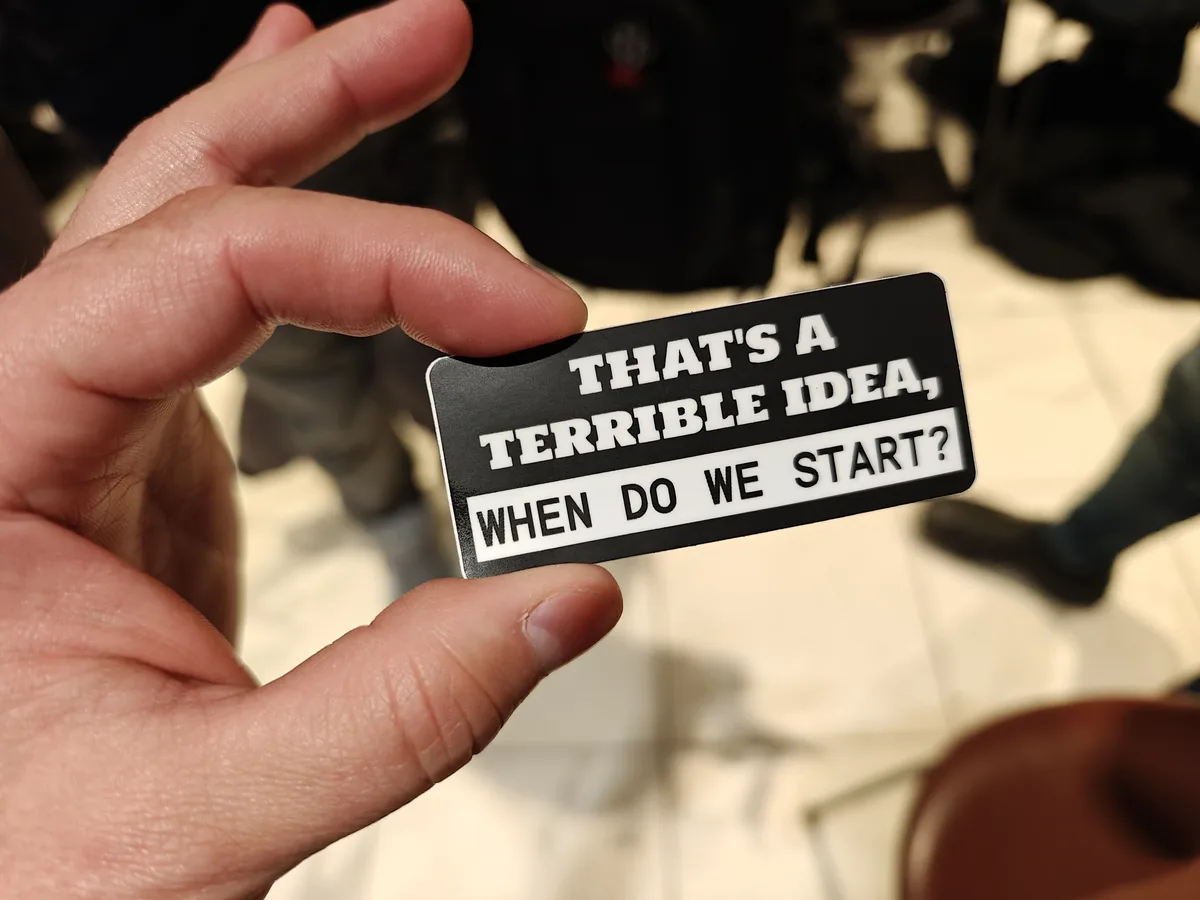 A hand holds a black rectangular sticker with white text that reads 'THAT'S A TERRIBLE IDEA, WHEN DO WE START?'. The…