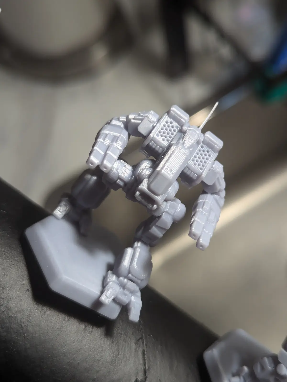 A close-up photo shows a small, unpainted, gray 3D-printed robot figurine standing on a hexagonal base. The robot has a…