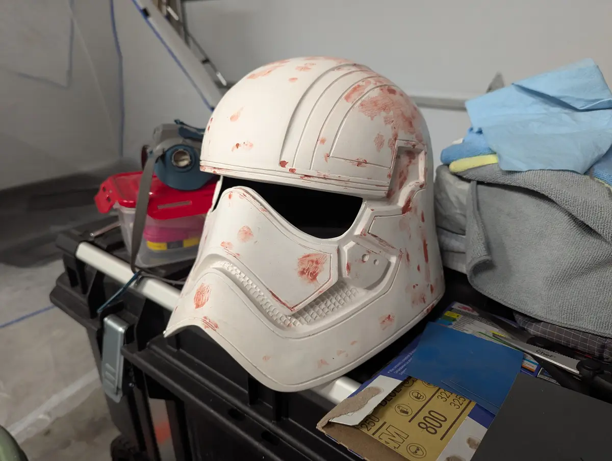 A white Star Wars-style helmet, likely a First Order Stormtrooper helmet, sits on a black toolbox covered in red paint…