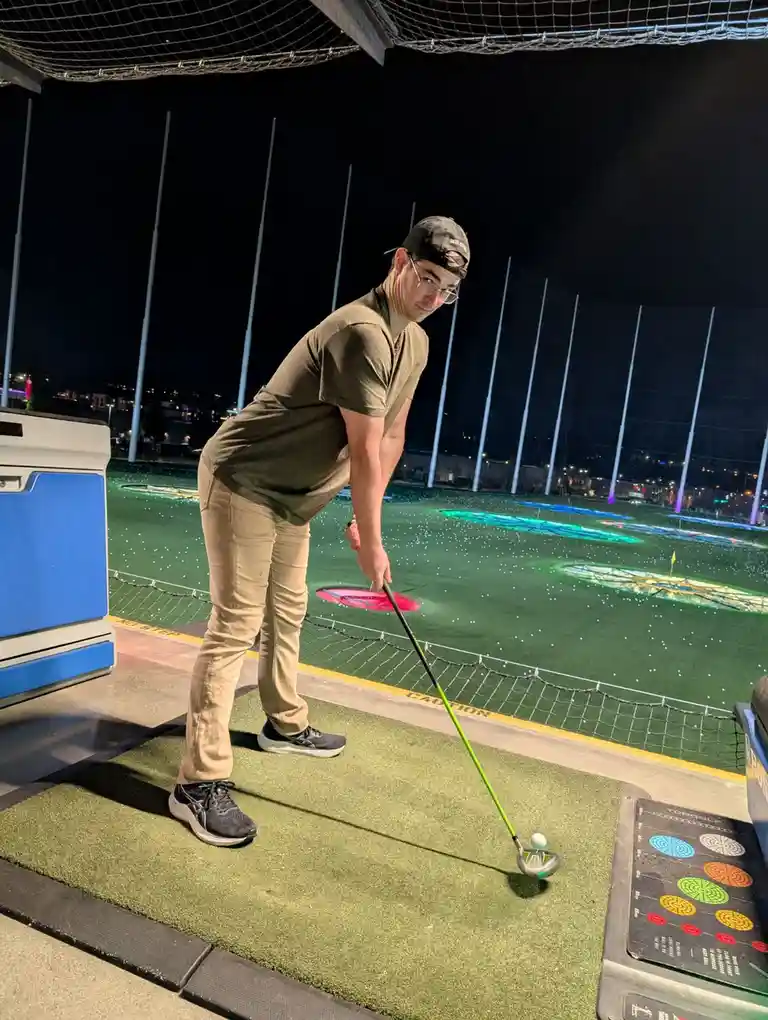A man in a dark t-shirt, beige pants, and a backward cap stands at a golf simulator bay at night, holding a golf club poised…